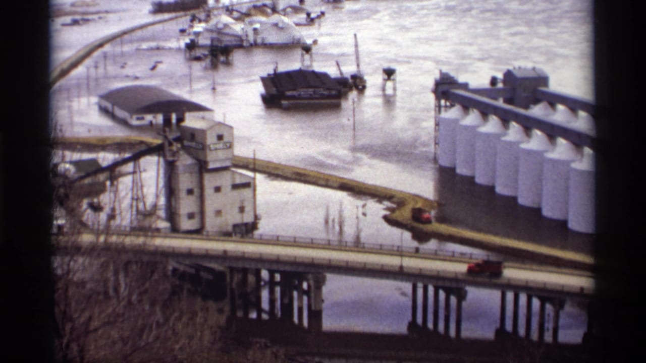 Flooded industrial riverfront with bridge, red car, grain elevator, and white silos.