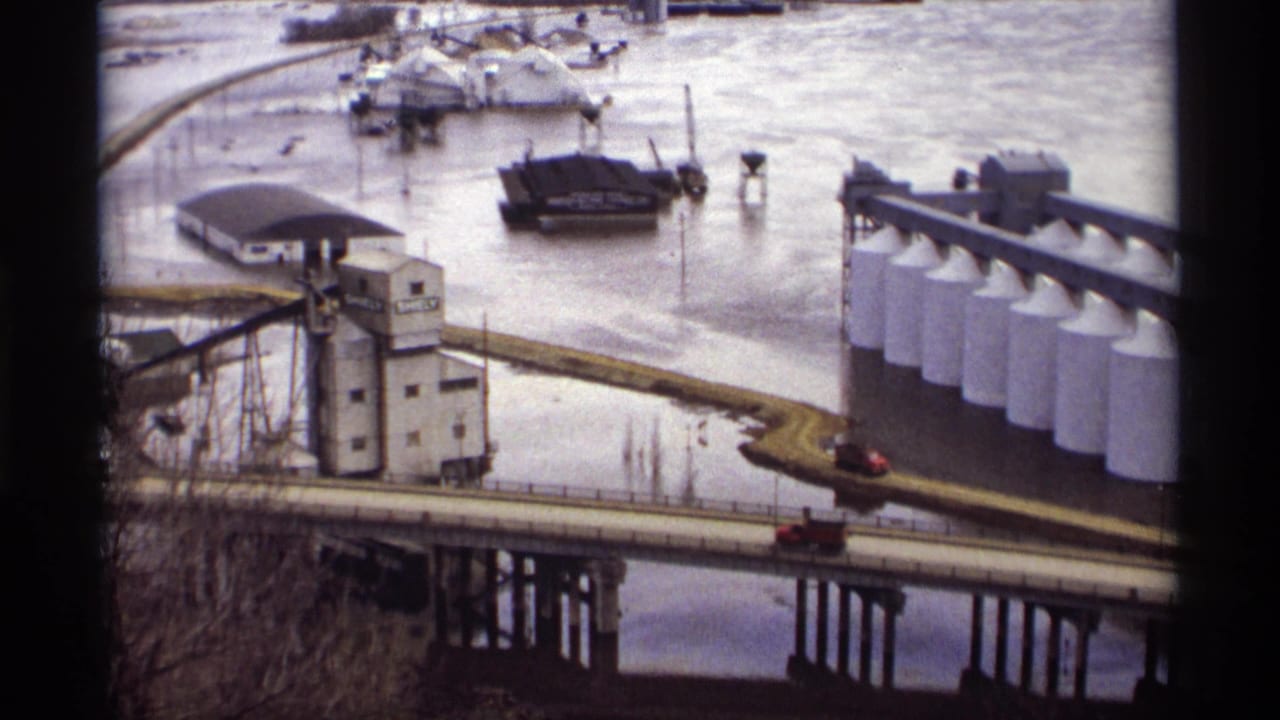 Bridge and flooded industrial riverfront with silos, buildings, and a small red car.