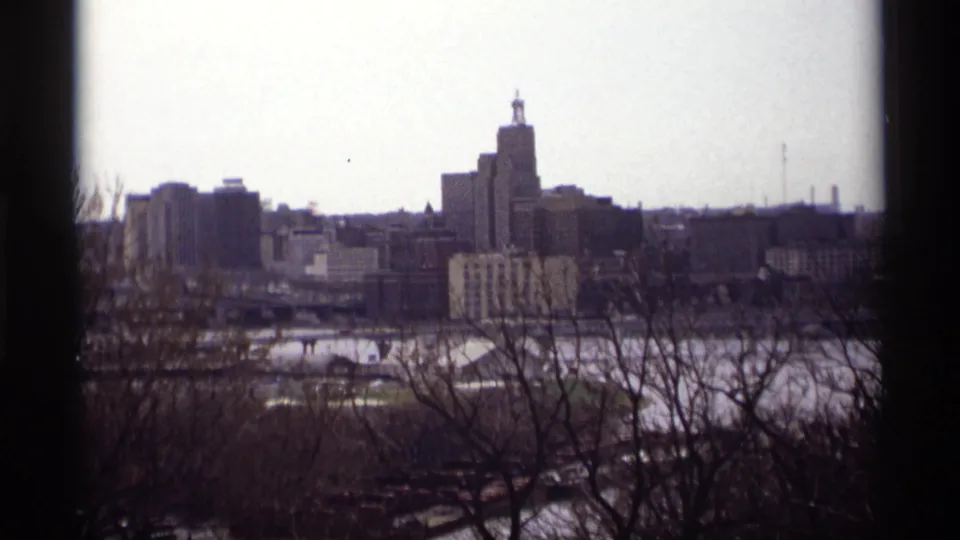 1969 Saint Paul skyline seen across water through bare tree branches, with downtown buildings under a pale sky.