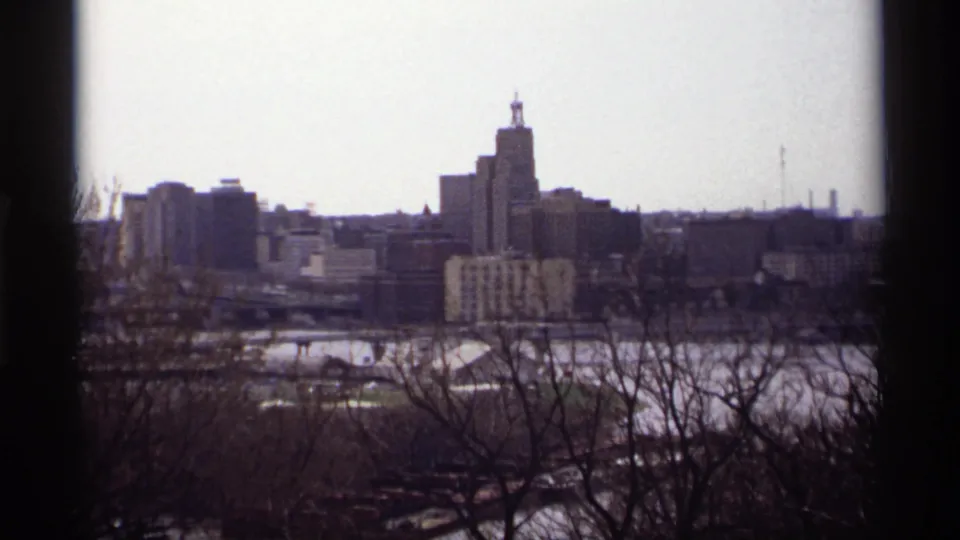 1969 Saint Paul 8mm skyline view with tall downtown buildings, leafless branches, and water below the overlook.
