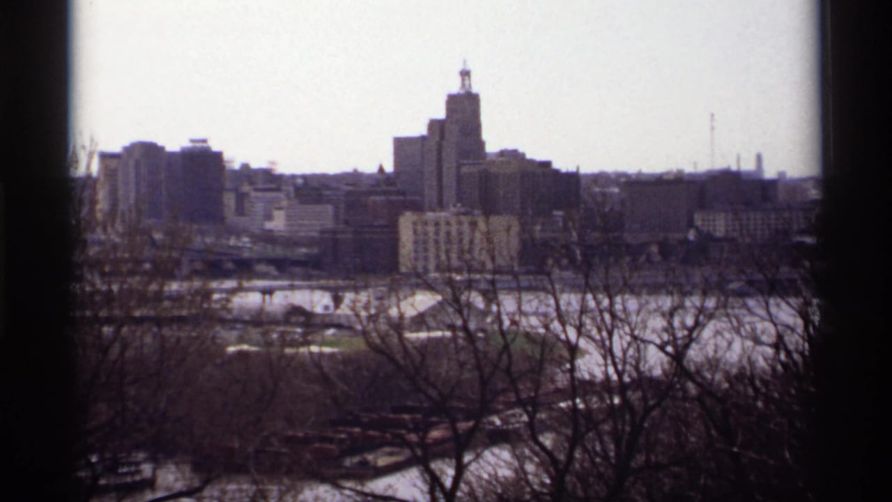 1969 8mm view of Saint Paul skyline with bare trees in the foreground and a tall building rising near the center.