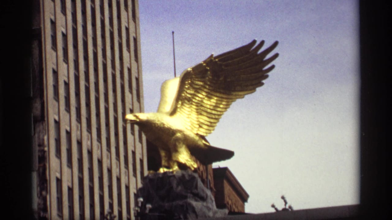 Gold eagle statue with spread wings beside a tall building and blue sky.