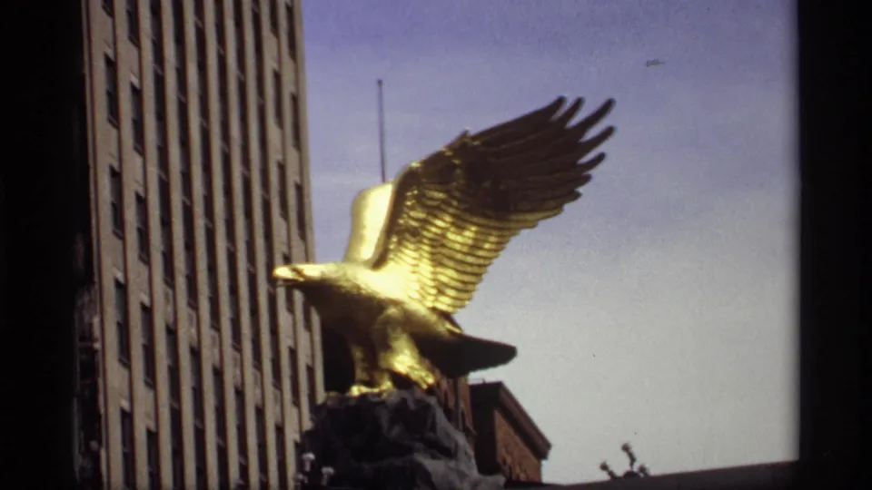 Closer 1969 Saint Paul frame of the gold eagle statue, its wing filling the sky beside the vertical lines of a tall building.