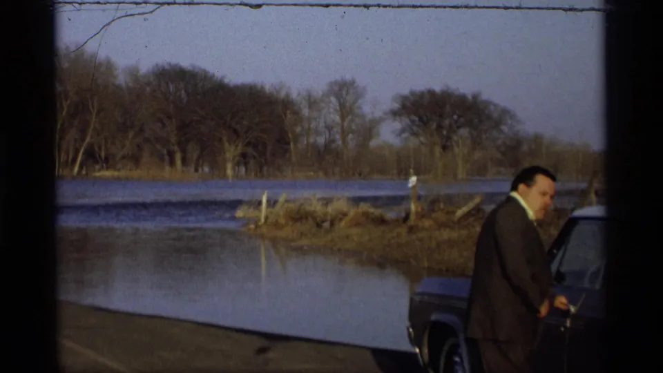 A man in a dark suit stands beside a car near roadside water and bare trees in the Savage, Minnesota home-movie sequence.