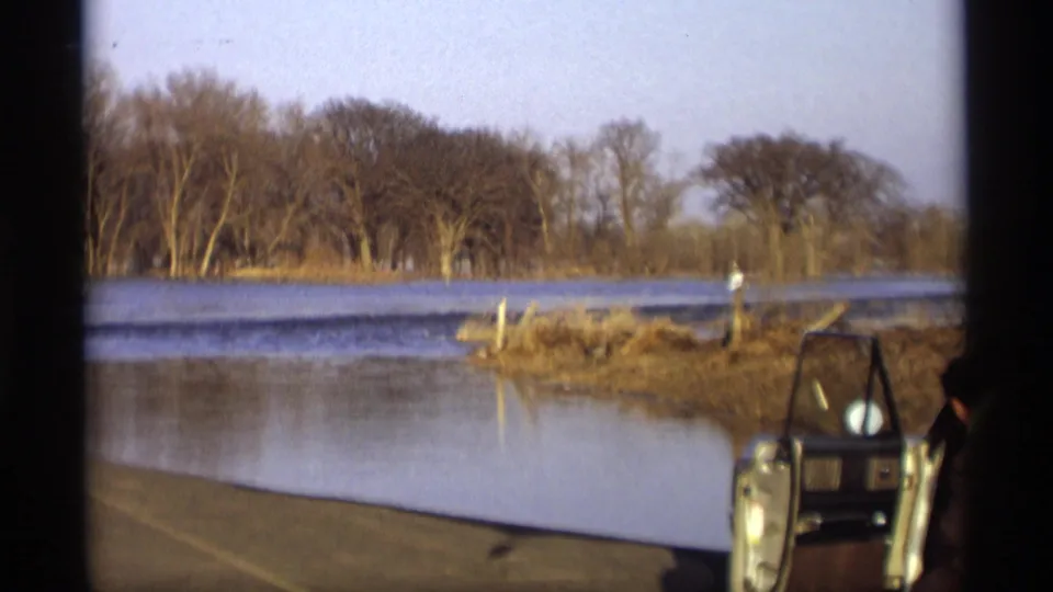 A classic car with its door open sits near shallow water in Savage, Minnesota, with bare trees and brush visible across the flooded area.