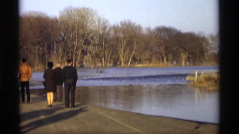 Three adults in coats stand on a road or park drive in 1969 Savage, Minnesota, watching water spread across the landscape.