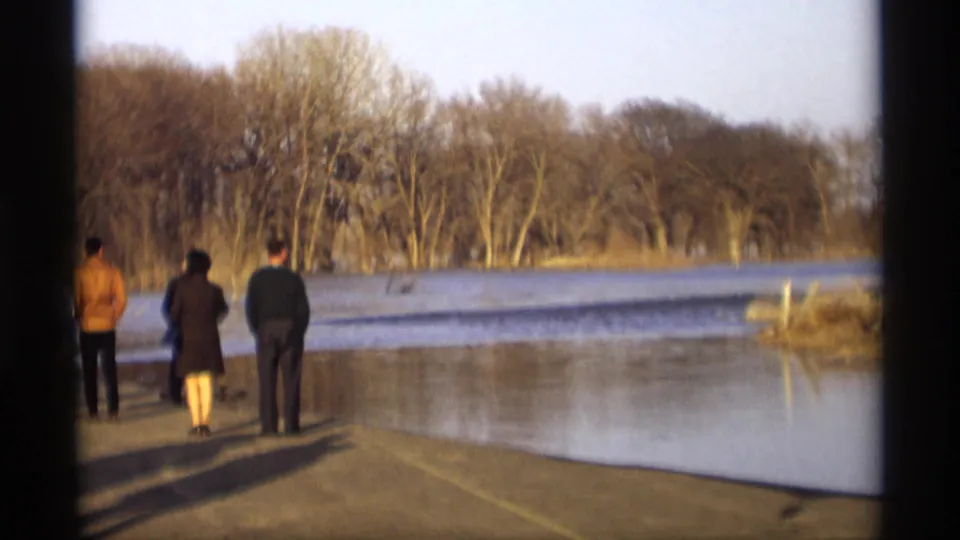 A small group of adults observes a flooded lakeside or park area in Savage, Minnesota, in a soft 1969 8mm home-movie frame.