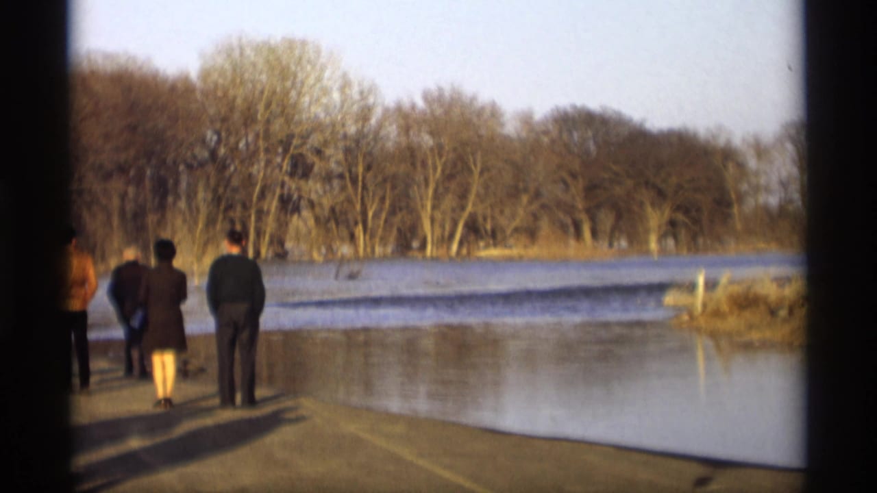 In Savage, Minnesota home-movie footage from 1969, adults in coats stand on a platform looking toward floodwater and winter-bare trees.