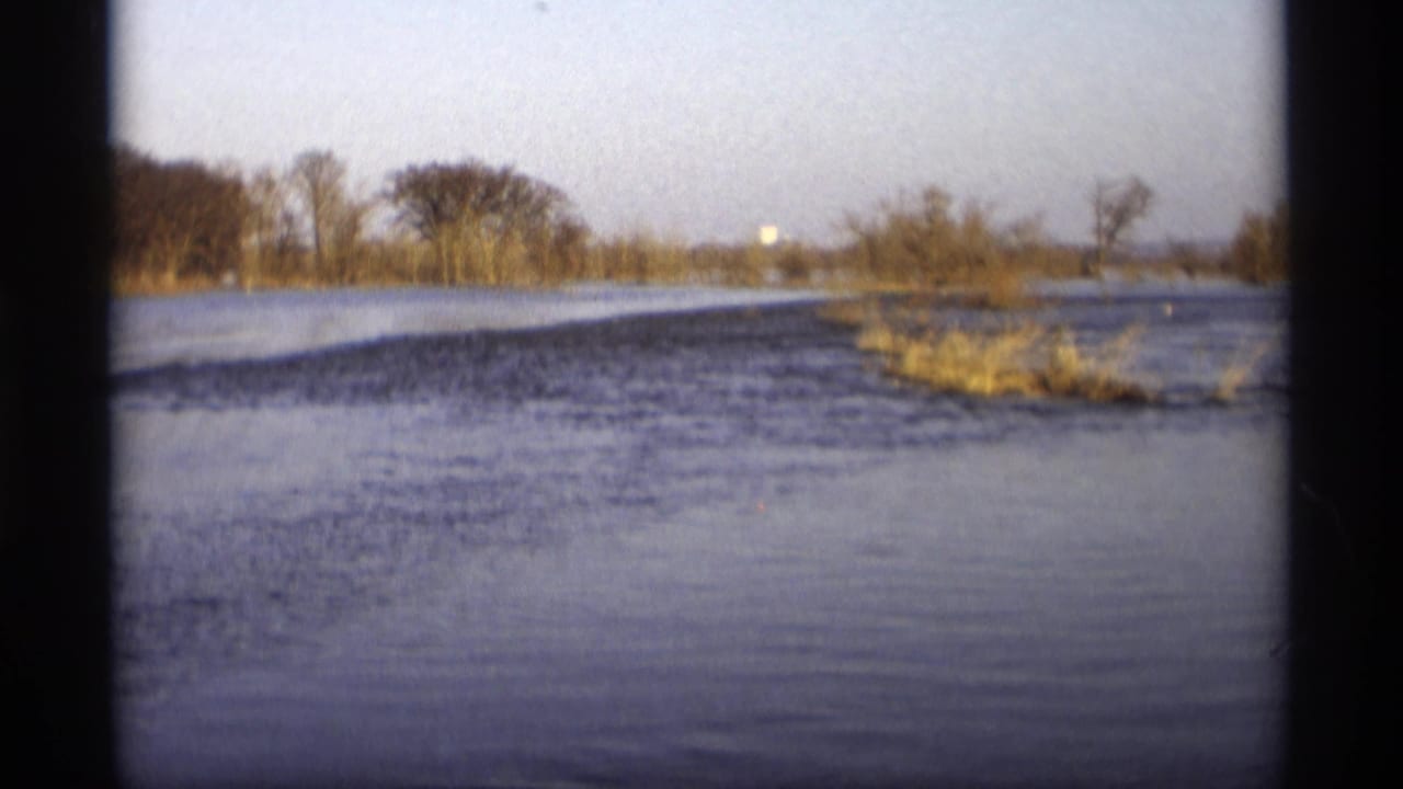 1969 Savage, Minnesota 8mm frame showing broad floodwater across a rural stream area with leafless trees and brush in the distance.