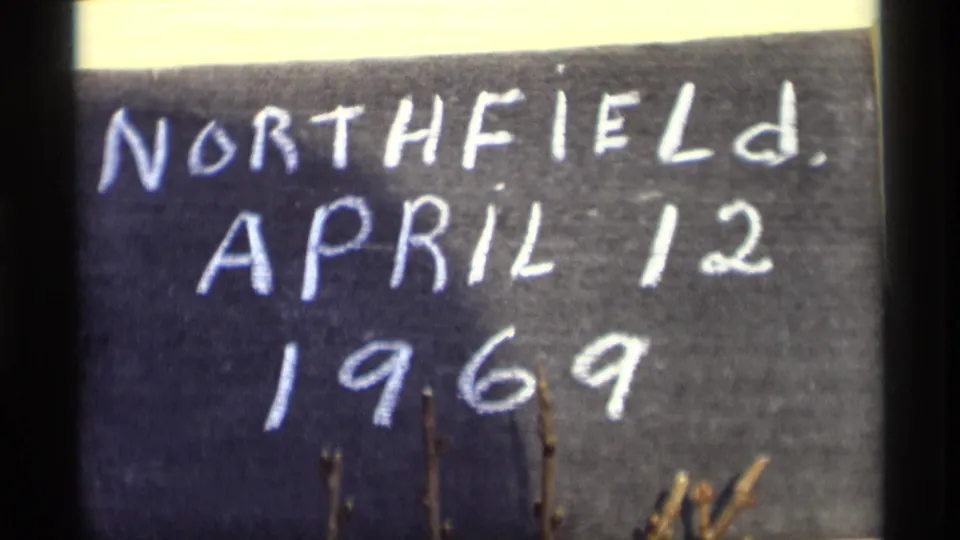 1969 home-movie title card showing Northfield, April 12, 1969 in white chalk, with warm film color and small branches in front.