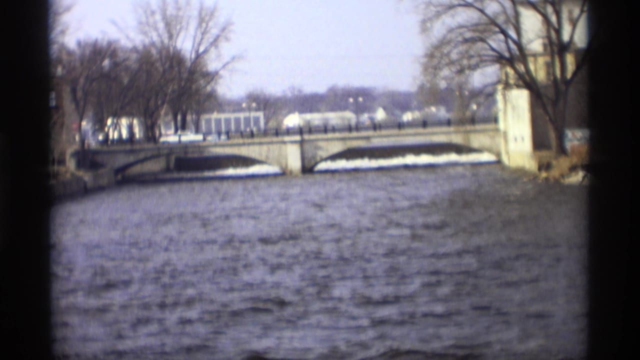 1969 Minnesota home-movie frame showing a choppy river, stone arch bridge, bare trees, and distant pale buildings near the far bank.