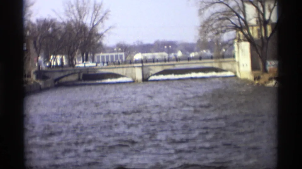 1969 Minnesota home-movie frame showing a choppy river, stone arch bridge, bare trees, and distant pale buildings near the far bank.