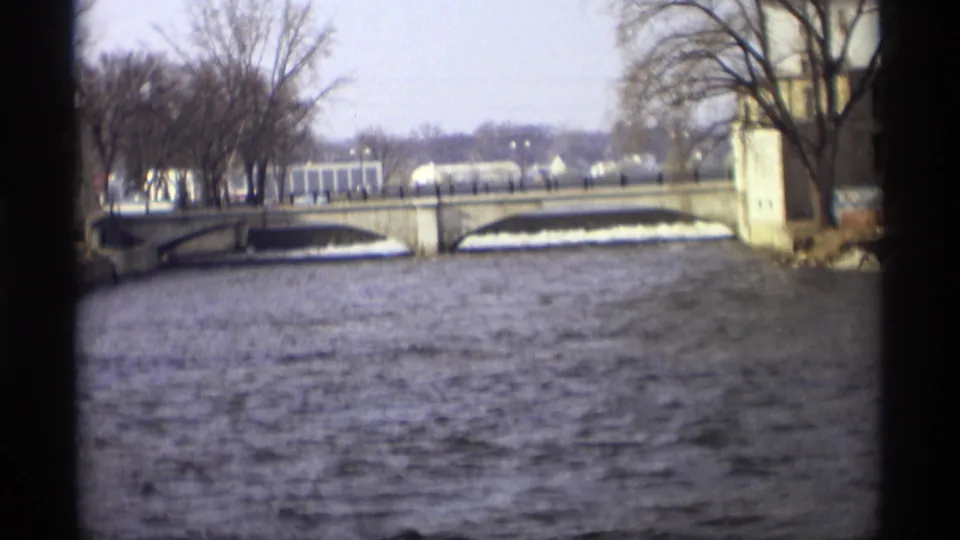 Late-1960s 8mm frame of the river surface leading toward a stone bridge, with leafless trees and distant structures visible beyond it.