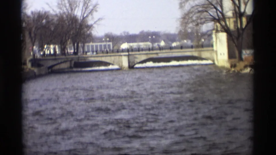 Northfield-area river view from the 1969 metadata, showing rippled water, a low bridge, bare riverbank trees, and pale structures in the distance.
