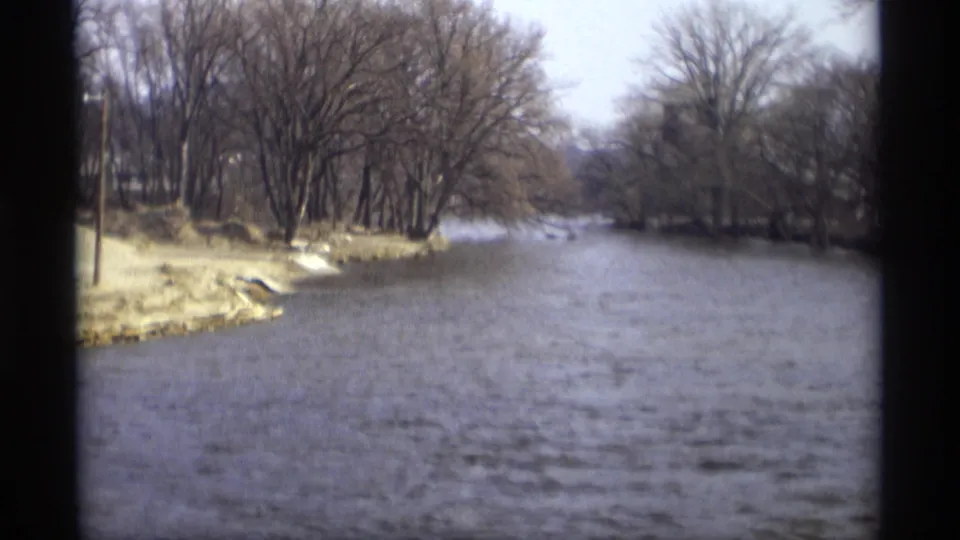 1969 home-movie frame of a wooded river bend in Savage, Minnesota, with bare trees and choppy water.