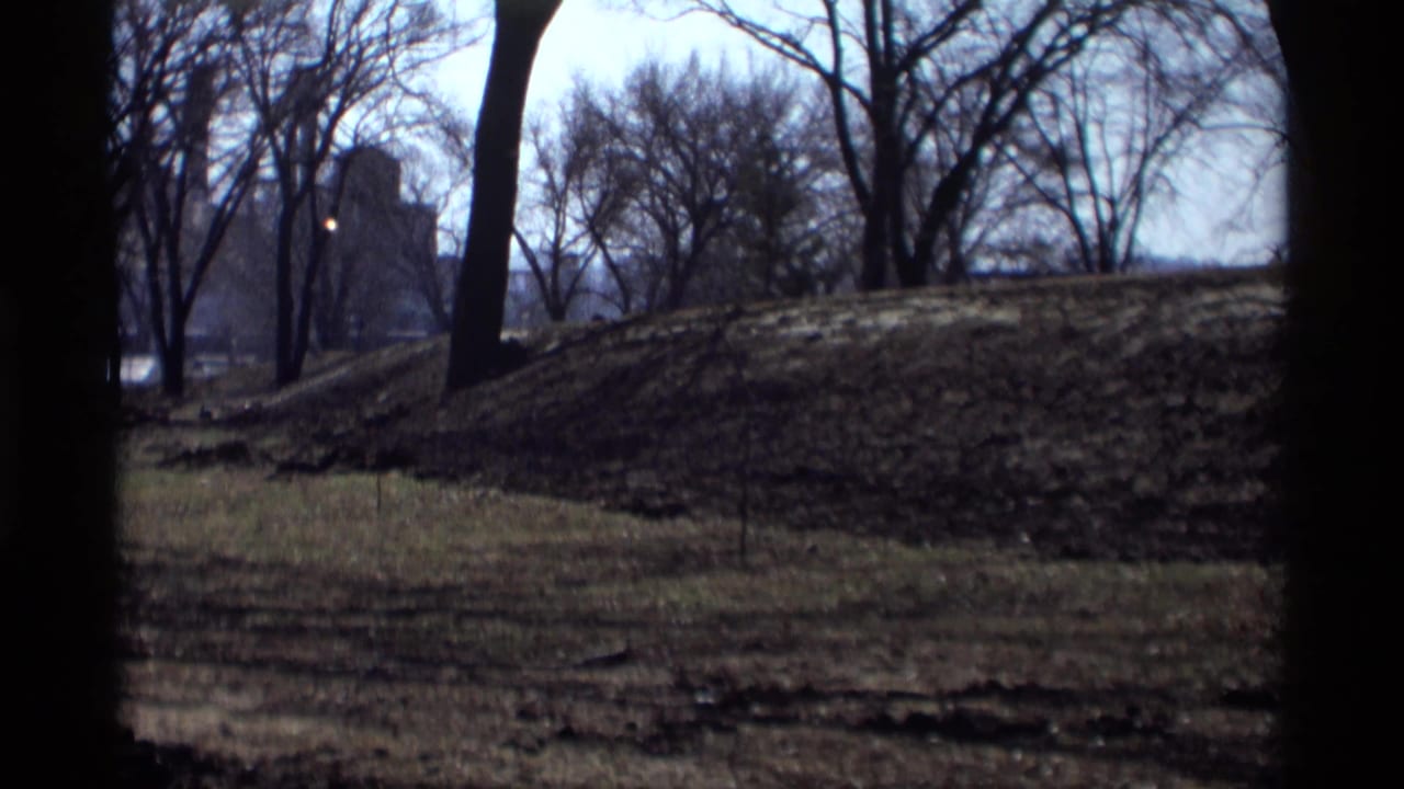 1969 8mm view in Savage, Minnesota, showing bare trees, patchy grass, a low mound, and a distant building beyond the park.