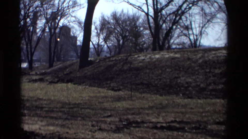 1969 8mm view in Savage, Minnesota, showing bare trees, patchy grass, a low mound, and a distant building beyond the park.