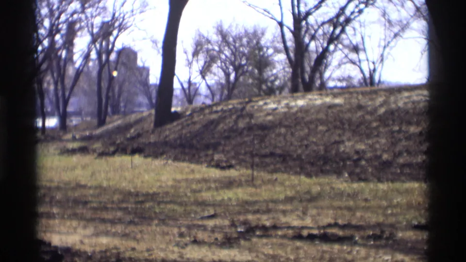 Vintage 8mm park view in Savage, Minnesota, with open grass, leafless trees, a low mound, and a distant building softened by focus and film grain.