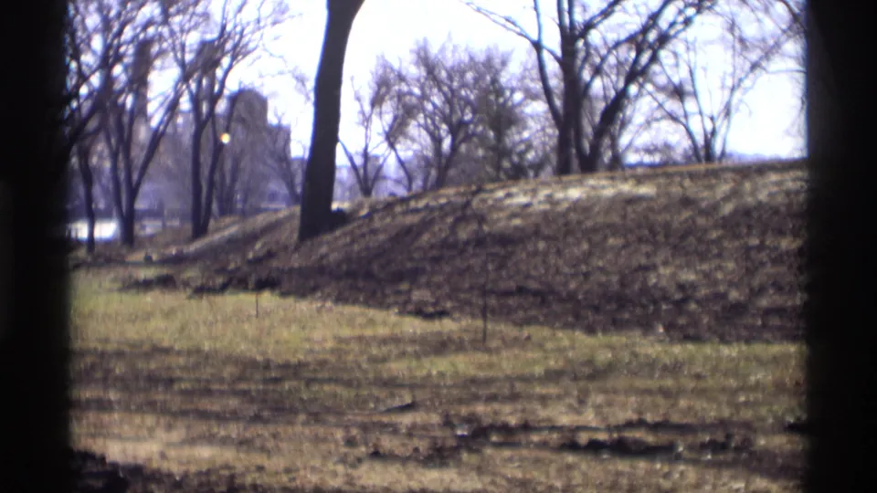Savage, Minnesota 1969 8mm frame showing a sunlit grassy mound, bare tree trunks, patchy ground, and a bright pale sky.