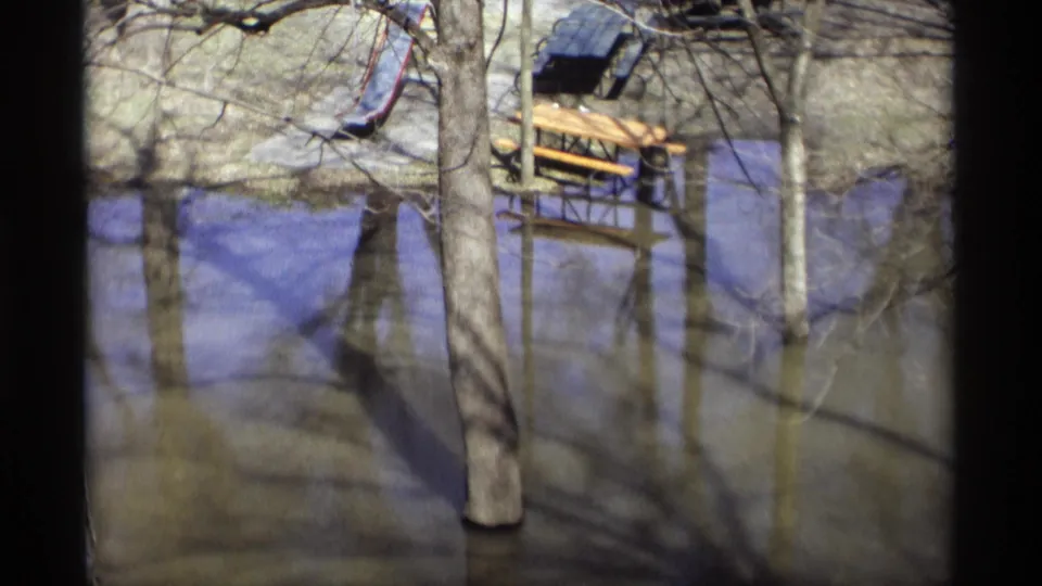 1969 8mm frame from Savage, Minnesota, showing floodwater around a picnic table, slide, and bare trees in a park.
