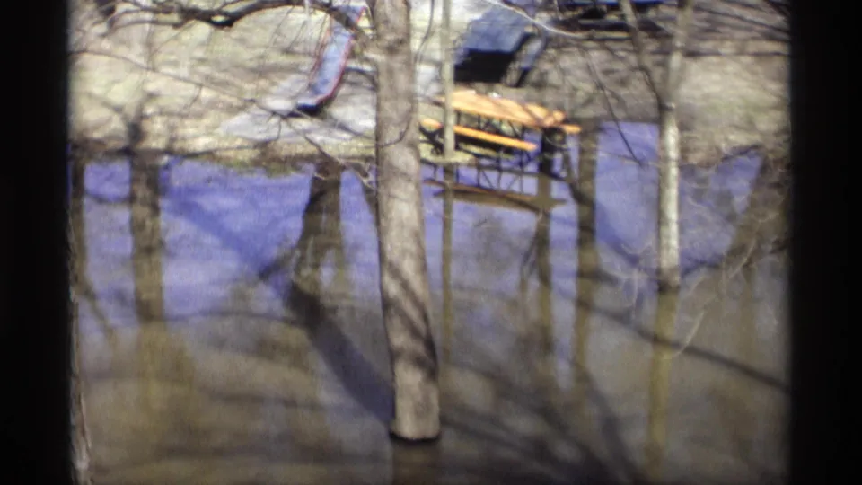 1969 8mm view of a Savage, Minnesota park flooded around bare trees, with a picnic table and slide visible behind them.