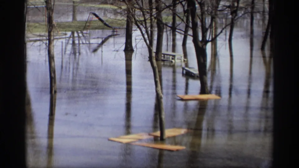1969 home-movie frame from Savage, Minnesota, looking across floodwater at bare trees, floating planks, a picnic table, and playground equipment.