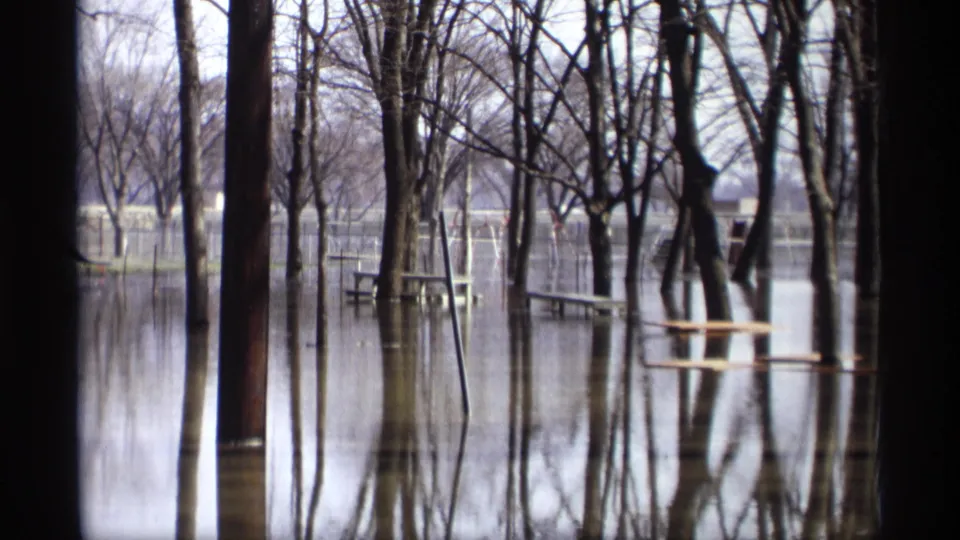 1969 home-movie view of a flooded Minnesota picnic area with tables, bare trees, and mirrored reflections on still water.