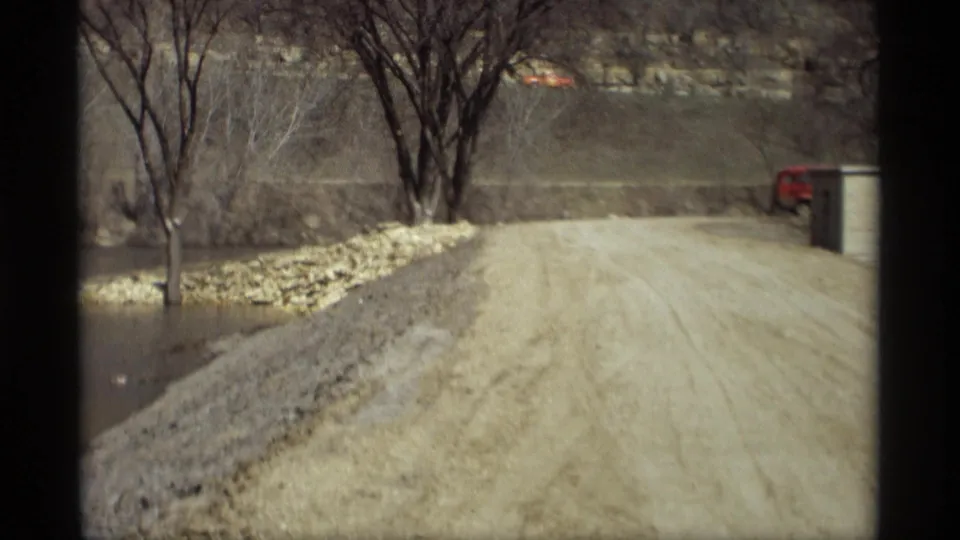 1969 Savage, Minnesota roadside scene with floodwater, bare trees, stones, and a distant red vehicle.