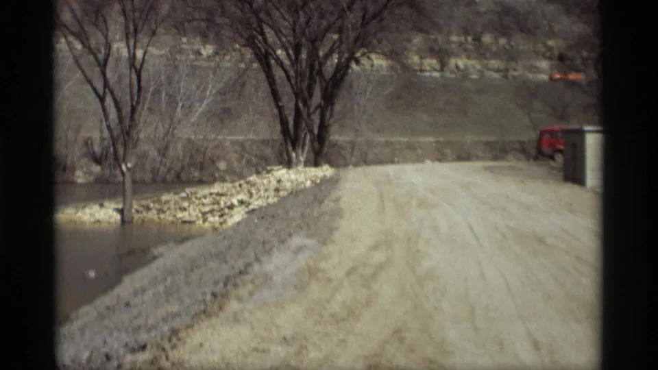 Savage, Minnesota 1969 home-movie frame showing a gravel road beside floodwater and a distant red vehicle.