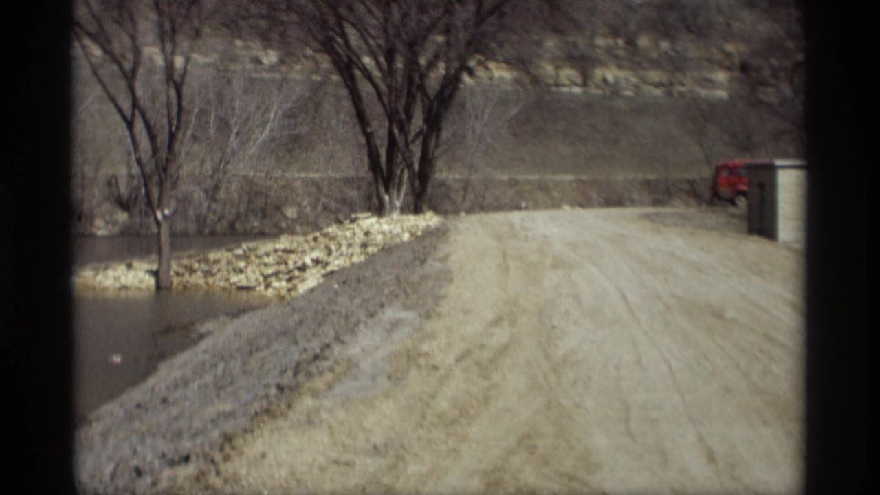 Dirt road beside floodwater with bare trees and a red vehicle near a small structure.