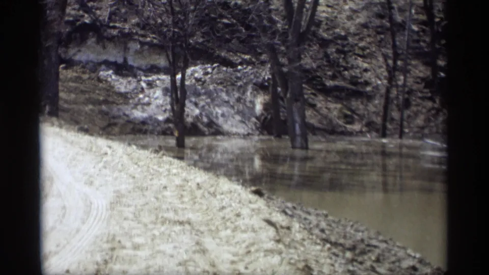 1969 Savage, Minnesota frame with a pale dirt road curving beside flooded trees and a rocky creek embankment.