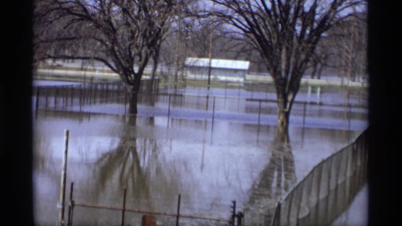 1969 8mm view from Savage, Minnesota showing floodwater around leafless park trees, a submerged fence line, and a distant small building.