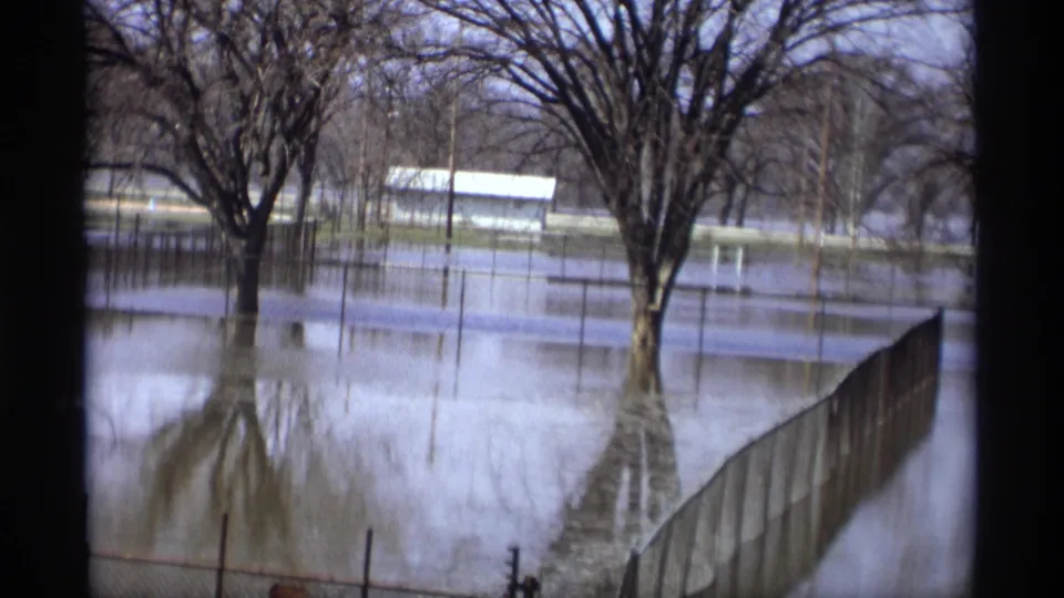 Savage, Minnesota flood footage from 1969 showing still water across a park, with bare trees, fence posts, and a small building in the background.