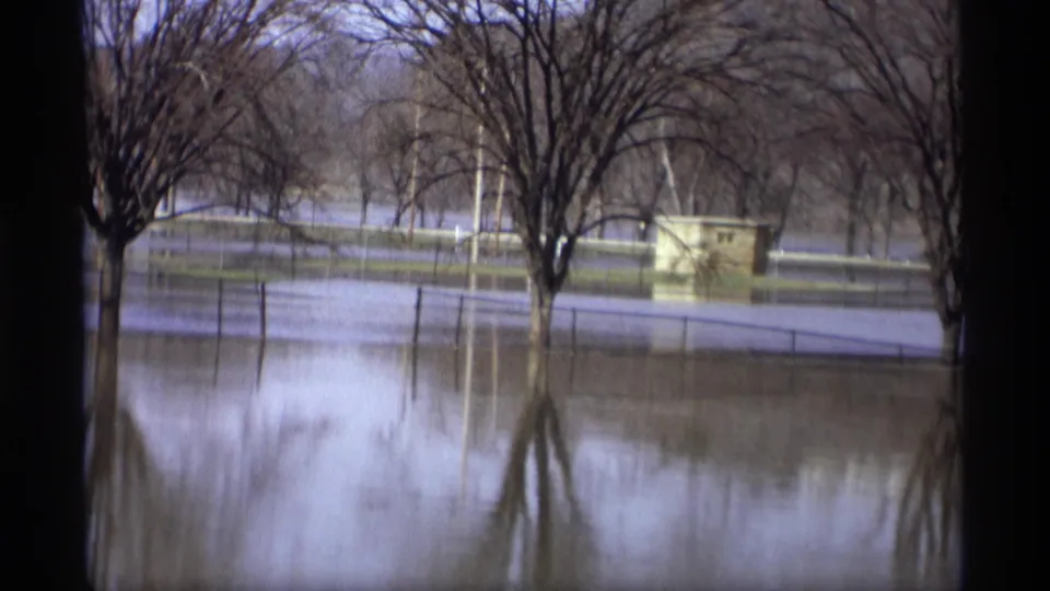 1969 Savage, Minnesota home-movie frame showing a flooded park area, bare trees reflected in the water, fencing, and a small structure across the water.