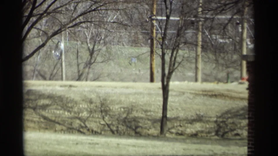 1969 Savage, Minnesota home-movie view through a window, showing leafless trees, pale grass, utility poles, and a brick wall edge.