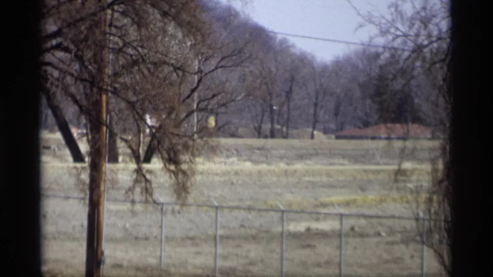 1969 Savage, Minnesota exterior with chain-link fence in the foreground, bare trees, a utility pole, open grass, and wooded hillside beyond.