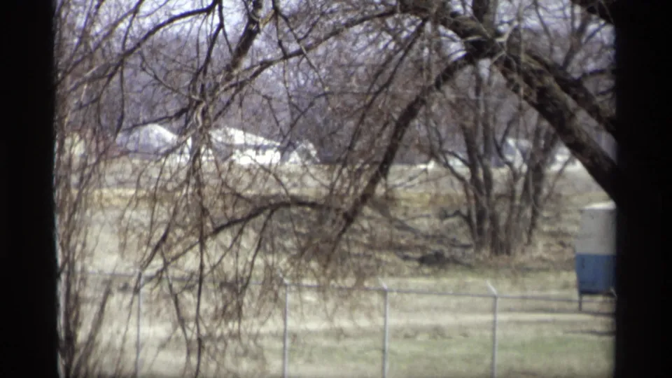 1969 Savage, Minnesota home-movie view through a window toward bare trees, pale grass, fencing, and distant low buildings.
