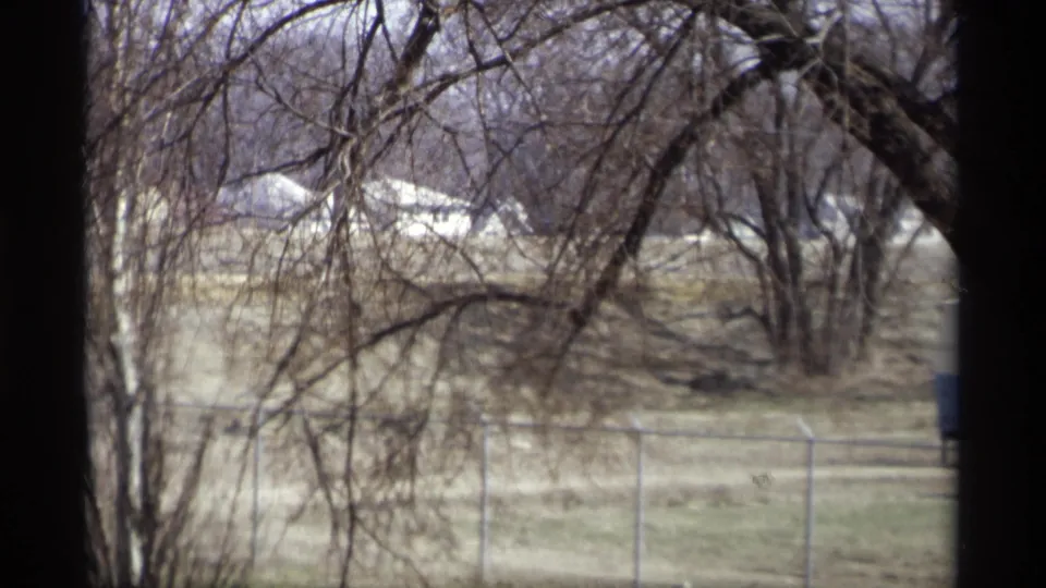 Window-framed 8mm view in Savage, Minnesota, showing bare trees, open field, chain-link fencing, and distant light-colored buildings.