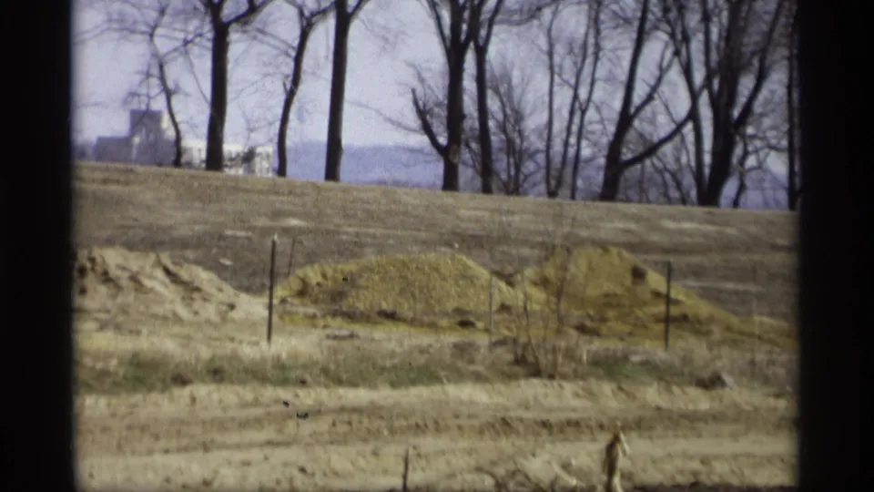 Savage, Minnesota 8mm frame from 1969 showing a muted field, sandy foreground, bare trees, and distant building forms near the horizon.