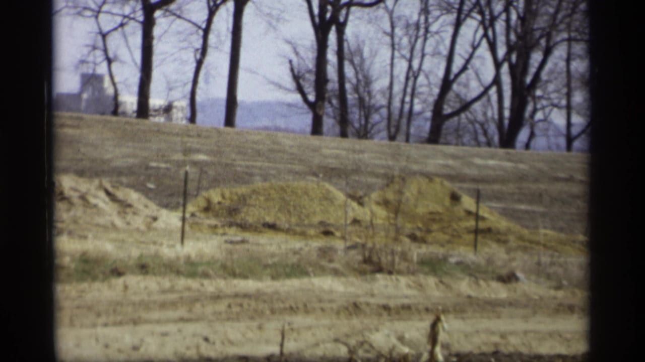 1969 8mm view in Savage, Minnesota showing a dry brown field, leafless trees, and a distant pale building partly hidden by branches.