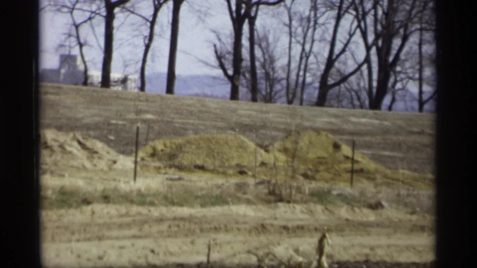 1969 8mm view in Savage, Minnesota showing a dry brown field, leafless trees, and a distant pale building partly hidden by branches.