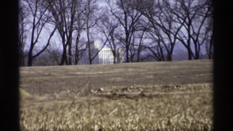 Savage, Minnesota landscape frame from 1969 with a harvested-looking brown field, bare trees, and a white building visible through the branches.