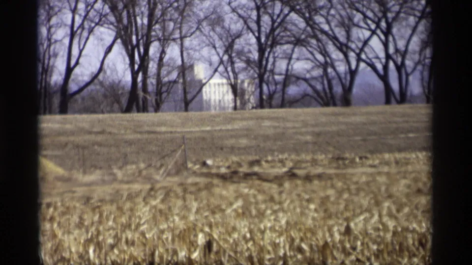 1969 home-movie frame from Savage, Minnesota showing stubbled field ground, bare trees, and a distant white building partly obscured by trunks.