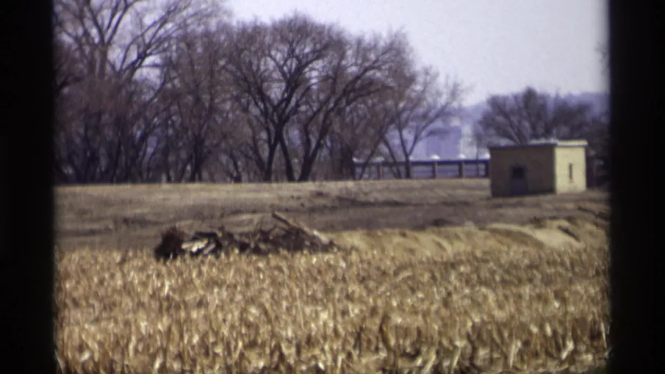 Final 1969 Savage, Minnesota frame shows a brown field, leafless trees, a small utility shed, a fence, and distant low hills or tree-covered ground.