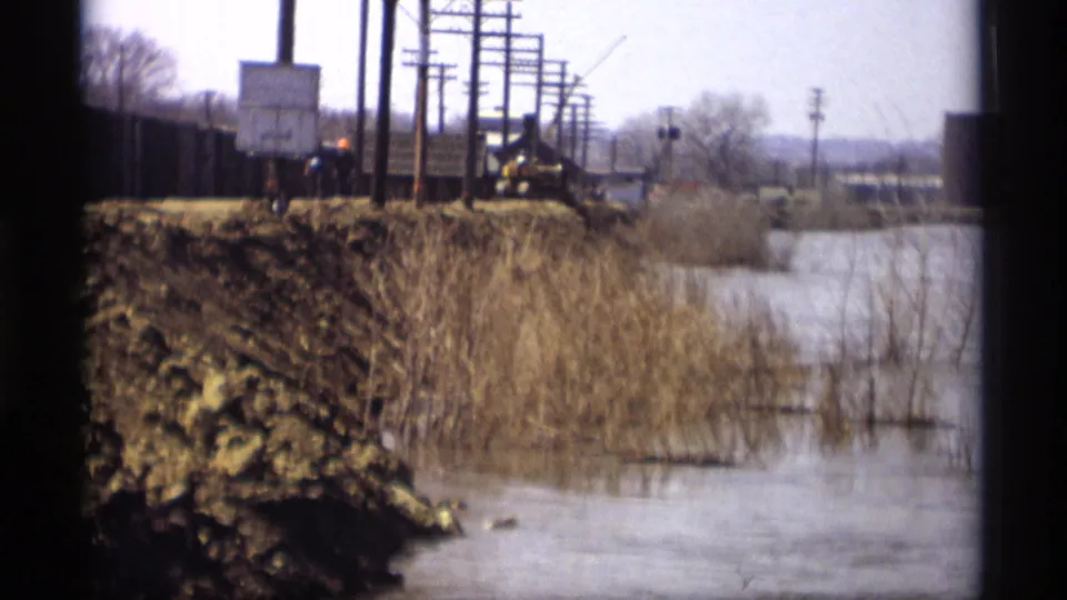 1969 Savage, Minnesota flooded ground beside a muddy embankment, with reeds, utility poles, and distant roadside structures.