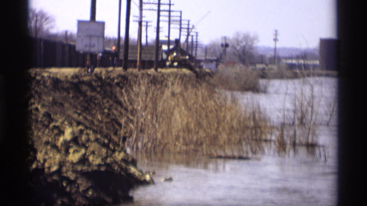 Savage, Minnesota 1969 frame showing a wet rail-side area, utility poles, muddy embankment, and distant rail equipment.