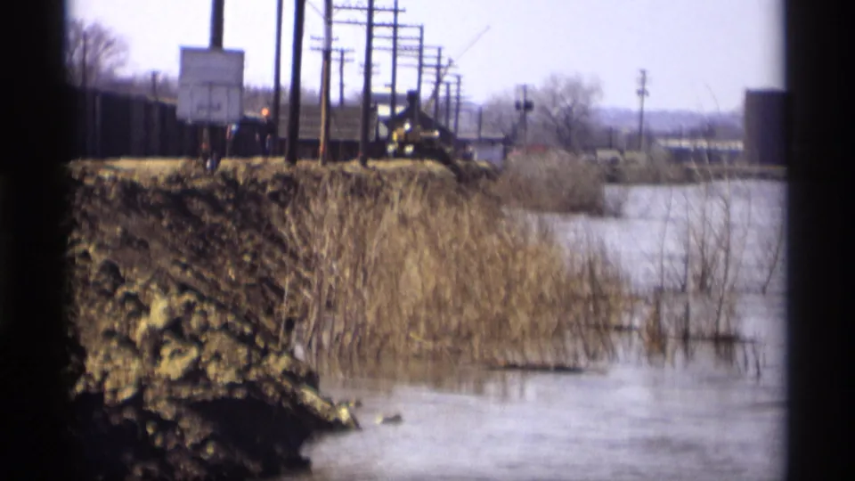 Savage, Minnesota 1969 frame showing a wet rail-side area, utility poles, muddy embankment, and distant rail equipment.