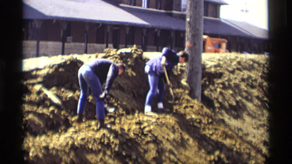Two workers dig with shovels on a muddy embankment near a utility pole in Savage, Minnesota, during the 1969 home-movie clip.