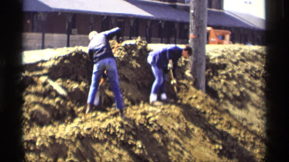 Savage, Minnesota 1969 frame of two workers using shovels on a muddy slope near a utility pole and rail-side building.