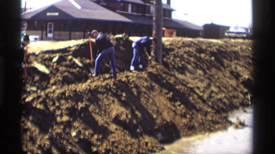 Wide 1969 Savage, Minnesota view of workers digging along a muddy trench near standing water and rail-side buildings.
